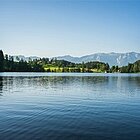 Blick auf einen ruhigen See mit blauem Wasser und blauem Himmel, Bäume und Berge im Hintergrund.